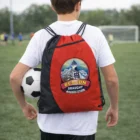 Boy with a soccer ball and Emblem Drawstring Bag on a field, players and goal behind.