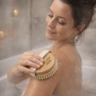 Woman using Hyacinthe Body Brush in a steamy bath, smiling as she brushes her shoulder.