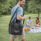 Man with black Innsbrook Cooler Bag; couple picnics in lush park background.