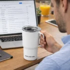 Man holding a Camelbak Horizon Vacuum Mug 700ml with YOUR LOGO HERE by a laptop.