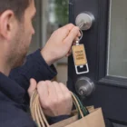 Person unlocks door, holds bags and a Santo Bottle Opener Metal Key Ring with YOUR LOGO HERE.