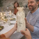Two people exchange Cotton Wine Bottle Gift Bags at a dinner table with food and candles.