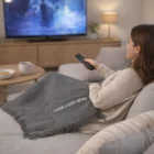 Woman on couch watching TV with a Polylana Acrylic Blend Throw Blanket labeled Your Logo Here.