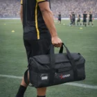 Person holding an Aquinas Water Repellent Duffle Bag on a soccer field with players practicing.