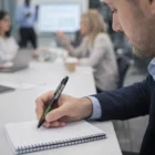 Man in suit writing with Paragon Aluminium Stylus Pen; blurred meeting colleagues behind him.