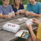 Four smiling people play with Paper Playing Cards With Gift Cases at a wooden picnic table outdoors.