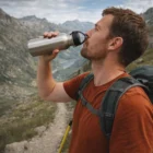 Man hiking drinks from Screw On Carry Lid Drink Bottle while wearing a red shirt.