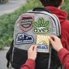 Someone adds a Polyester Woven Patch to a gray backpack with other patches.