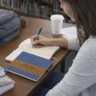 Woman writes in a Denim Finish Notebook at a table with coffee and her backpack nearby.