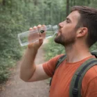 Man drinks from 600Ml Glass Bottles on a forest trail, wearing an orange shirt and backpack.