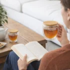 Person reading, holding a 250Ml Glass Sets mug of tea with bamboo lid near a teapot.
