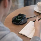 Someone writes in a notebook while a phone charges on a 15W Wireless Charger on a round table.