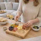 Woman arranges cheese, fruit, and meats on a Square Cheese Board with wine and snacks nearby.