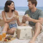 A smiling couple enjoys sandwiches and drinks on the beach with 6L Jute Cooler Bags.