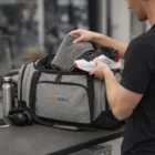 Man packs a Carter Duffle Bag with a towel and sneakers, next to headphones and water bottle.