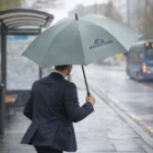 Man with Business Umbrellas With Logo Branding waiting at a rainy bus stop.