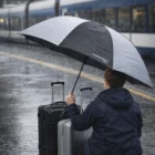 Traveler with suitcases holds a Hurricane Sport Amara Umbrella in rain near a train platform.