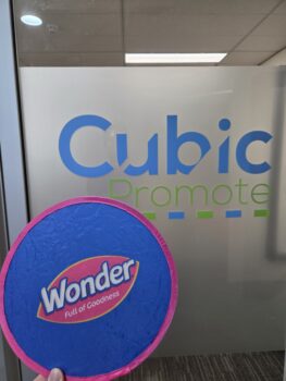 A hand holds a blue Wonder frisbee with the best logo format by a Cubic Promote office sign.