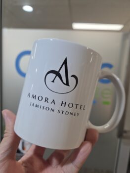 A white mug with Amora Hotel Jamison Sydney logo in crisp EPS format held in someone's hand.