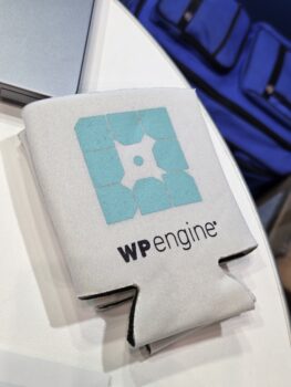 A white WP Engine koozie on a table, showing off the best logo format by a blue bag.