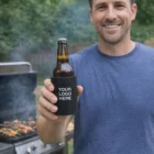 Smiling man holds a beer in Stubby Holders by a grill at an outdoor barbecue.