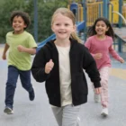 Three smiling children in Kids Relax Zip Hoodies run toward the camera on a playground.