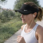 Woman jogging outdoors, wearing Nike Dri-Fit Swoosh Visor and earphones on a sunny day.