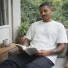 Man with tattoos reads on a patio, wearing Classic Organic Tees.