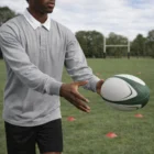 Person wearing Rugby Jersey passes a ball on grass field with goalposts in background.