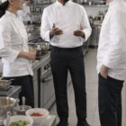 Three chefs in Spicemaster Jogger Pants chat and smile together in a commercial kitchen.