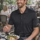 Smiling man in a Nike Dri-Fit Legacy Polo enjoys salad at an outdoor café on a sunny day.