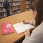 Woman writing in a Beautone Fashion Diary at a library table with a pink journal.