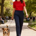 Woman in red shirt and Secret Waist Fit Pants walks a dog in a sunny park with trees nearby.