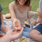 Woman shares Hinged Tin Jelly Beans For Expos at a picnic on a checkered blanket.