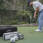 Man placing golf ball on green with Golf Balls Callaway and boxes displayed in the foreground.