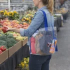 Woman with an apple at a market, carrying a Maddy Clear Tote Bag with blue handles.