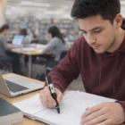 Young man writing in a library with books and Corporate Branded Flash Drives on the table.