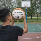 A person aims a Basketballs ball with YOUR LOGO HERE at an outdoor hoop.