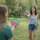 Two women play catch with Grippy Sticky Balls and paddles in a grassy park.