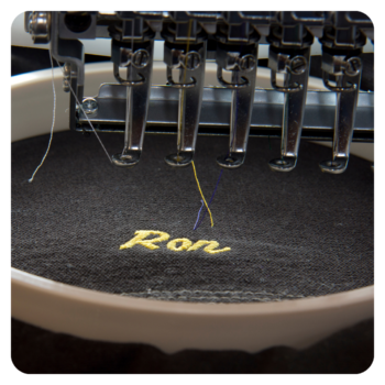 Close-up of an embroidery machine stitching "Ron" on black fabric for durable golf shirts.