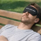 Man in gray shirt on bench wearing an Eye Mask that says YOUR LOGO HERE.
