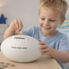 Smiling boy puts a coin into a Football Coin Savings Bank on the table.