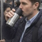 Man in a blue jacket uses an Alba Logo Branded Travel Mug on a crowded subway train.