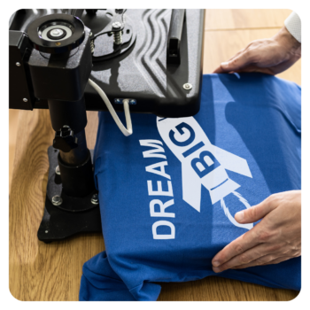 Hands pressing a blue golf shirt with DREAM BIG and a rocket using a heat press machine.
