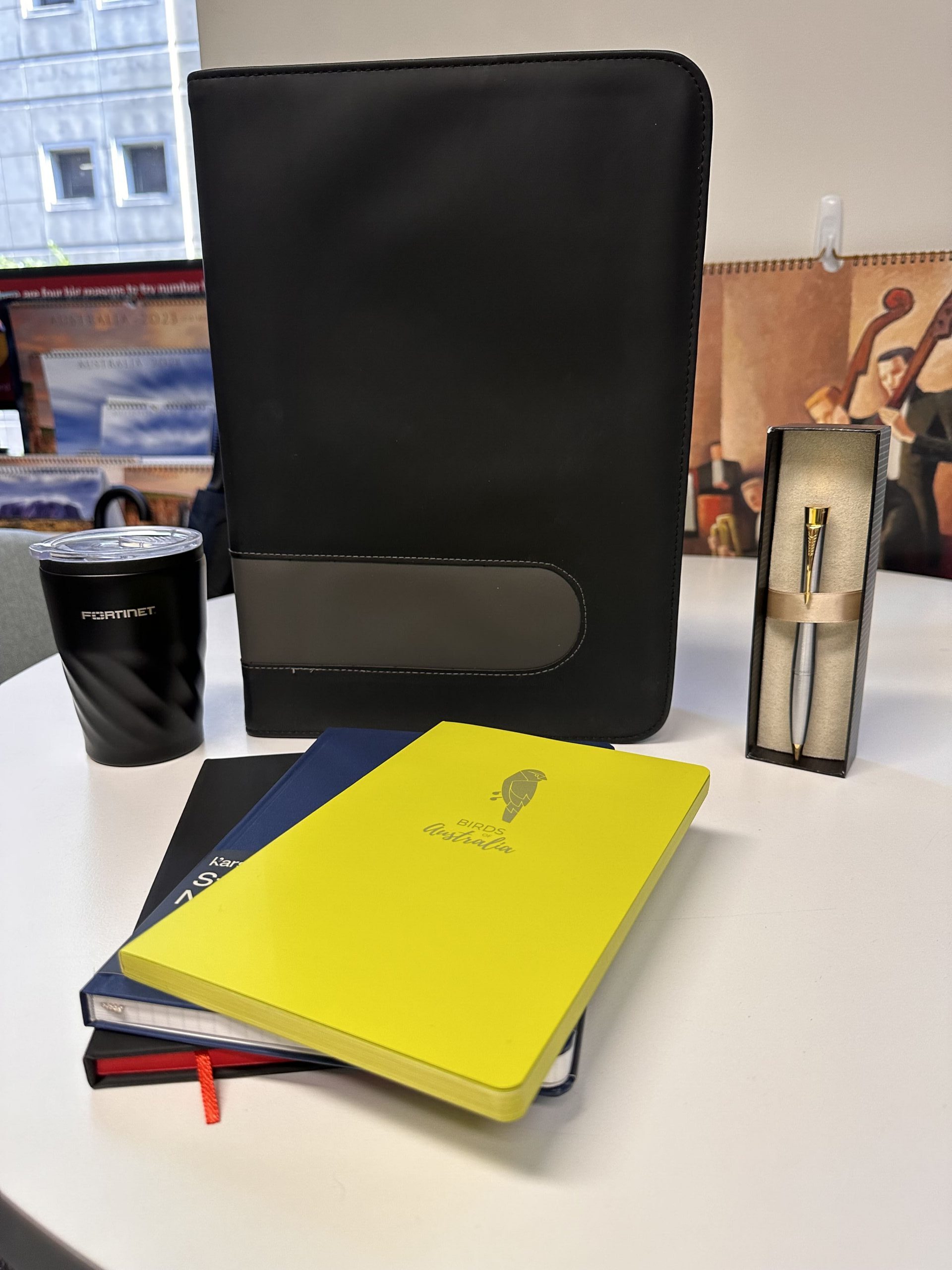 A branded merchandise forecast set: black folder, yellow notebook, pen set, and cup on a white desk.