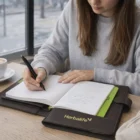 Woman writing in a notebook with coffee, phone, and an Ezra A5 Portfolio on the table.