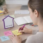 Woman writing notes at a desk with Cut Adhesive Notes and a house-shaped sign.