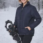 Smiling woman wears Pos Adult's Heavy Quilted Jacket, holding a snowshoe on snowy forest path.
