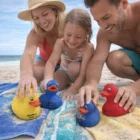 Smiling family playing with Quack Bath Ducks on a sandy beach, blue ocean in background.