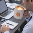 Man holding a Bistro Wheat Fibre Mug with Geek Shop logo, seated at a desk with notebook and laptop.